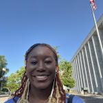 Simi Olabode, Baker-Nord Graduate Fellow: African American woman in blue shirt standing outside of a professional building with a displayed American Flag
