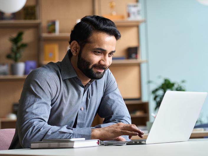 bearded man in a shirt sits at a desk, focused on his laptop.