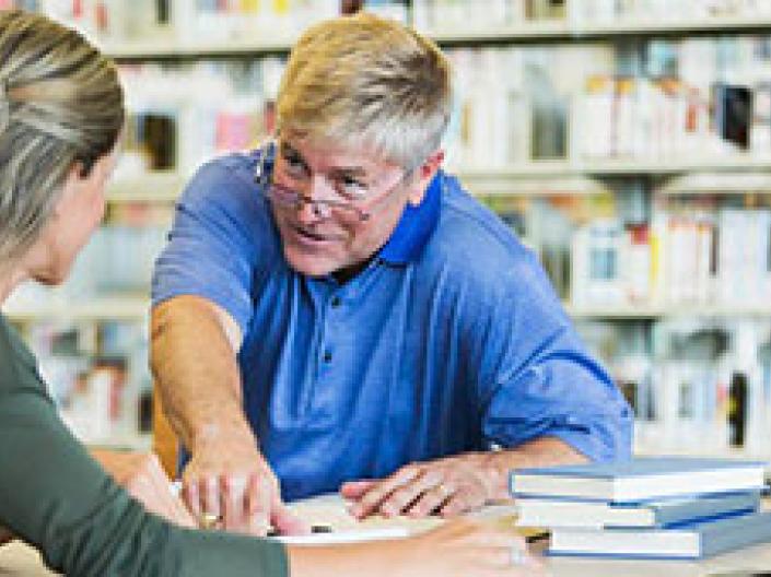 This is an image of a male professor helping a female student with her work while pointing to the paper on the table in front of them.