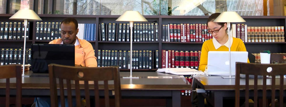 two students stuydying in the library on laptops, on a study table, with a row of books in background