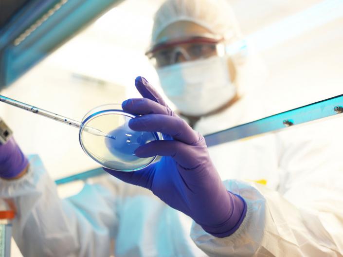 Person wearing a lab coat and face mask working in a lab with a petri dish