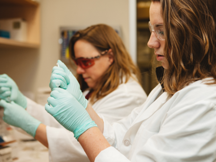 Two women in white lab coats and green sterile gloves work next to each other at a lab bench