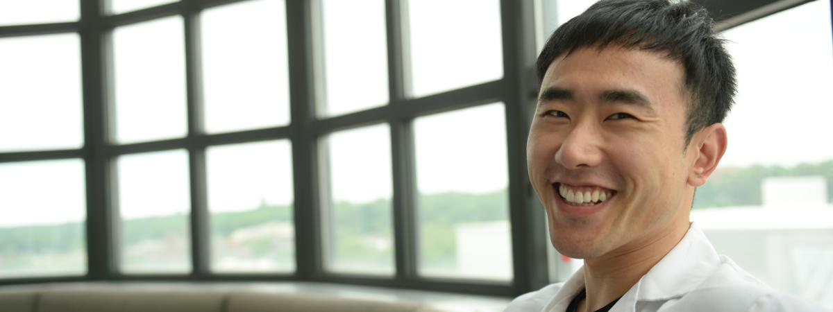 A smiling student wearing a white lab coat sits on a couch in front of a large window.