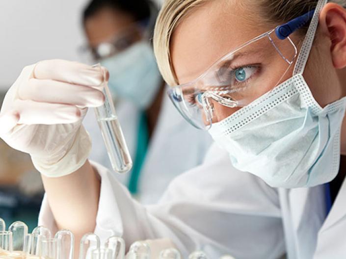 Image of lab professional with safty glasses, surgical mask, white lab coat and white latex gloves, analysing a clear test tube with a clear liquid, with test tubes in a rack, some upside down, in front of her. There is another lab professional dressed similarly in the background and a microscope in the midground to the left.