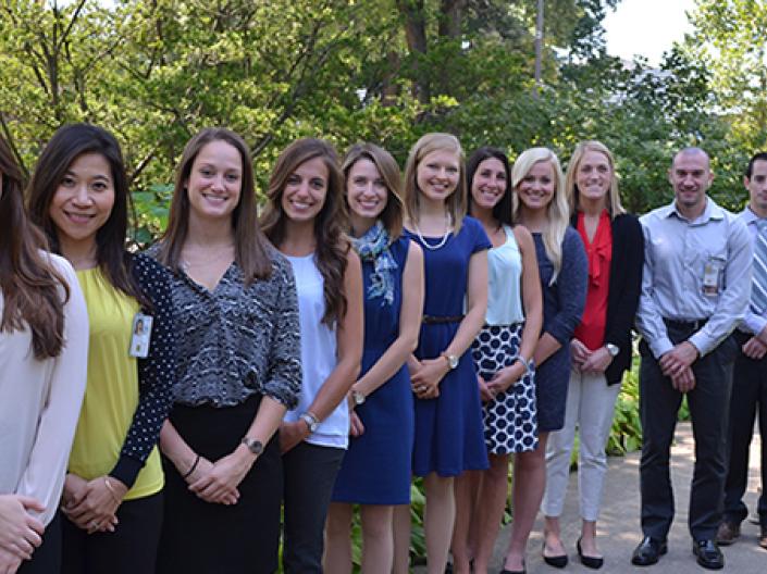 Image of twelve students in dress attire lines up in a row decending away from the camera smiling.  they are on a concrete path in a green wooded area with a large tree behind the first person on the left.  The sun is shining.