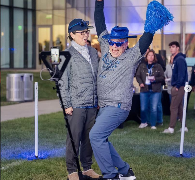 Two CWRU parents pose in a 360 photo booth with blue light up sunglasses, a blue cowboy hat and a pompom during Blue Bash.