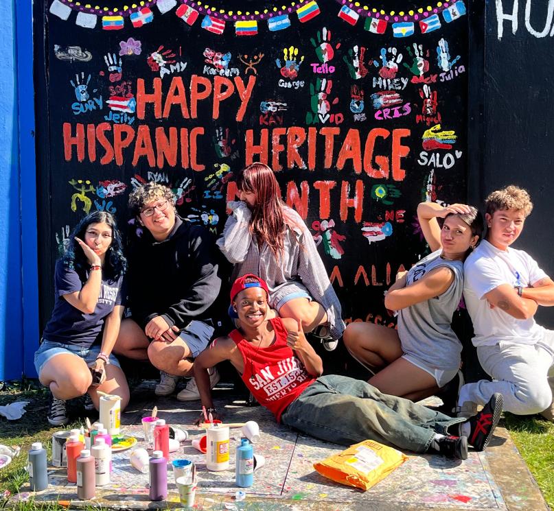 A group of six students pose in front of the CWRU spirit wall painted to say “Happy Hispanic Heritage Month.”