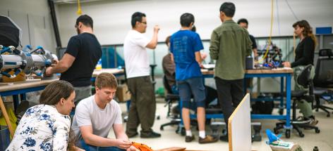 Two people examine a robotic crab on sand, with several other lab workers doing other activities in the background.