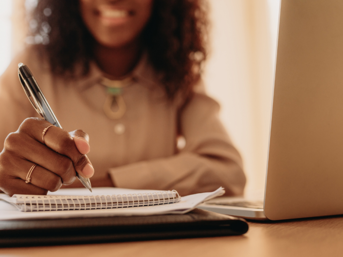 A woman rights in a notebook with her laptop open on her desk