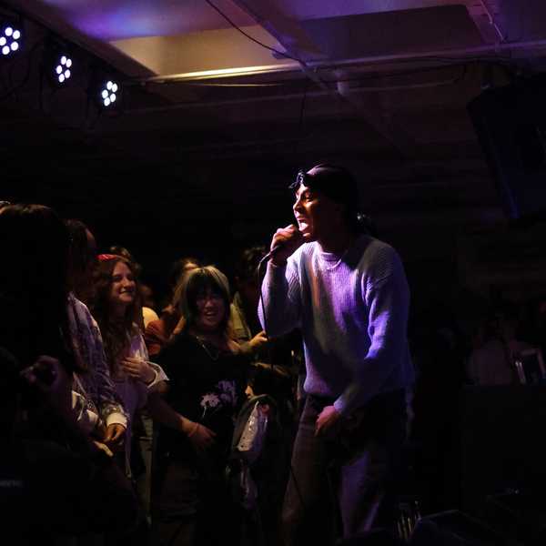 A student performer sings into a microphone under purple stage lights in a small, crowded venue, with audience members gathered closely.