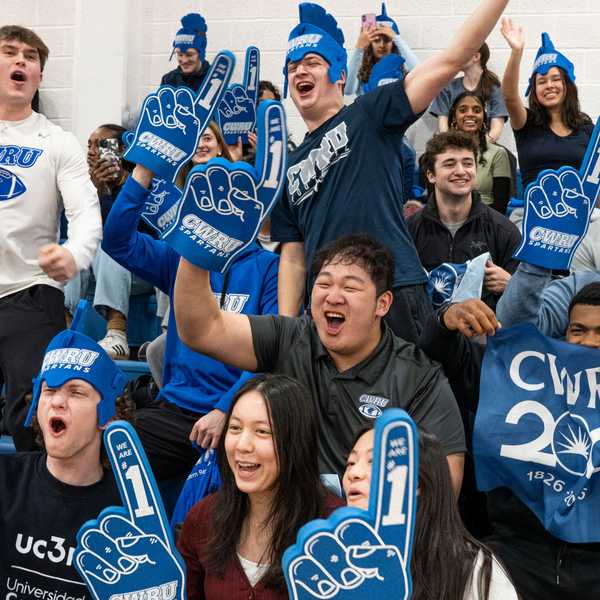 A large group of CWRU students wearing blue foam Spartan helmets and blue foam fingers, cheering on a basketball game at Horsburgh Gymnasium.
