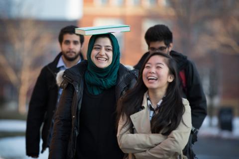 Students walking and laughing, one balancing a book on her head.