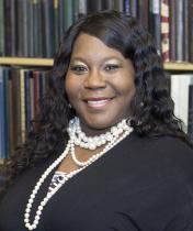 Angela Carpenter smiles at the camera in front of a bookshelf. She is wearing a black wrap top and a pearl necklace. 