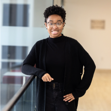 LaNyah, wearing glasses and an all-black outfit, leans against a railing and smiles at the camera