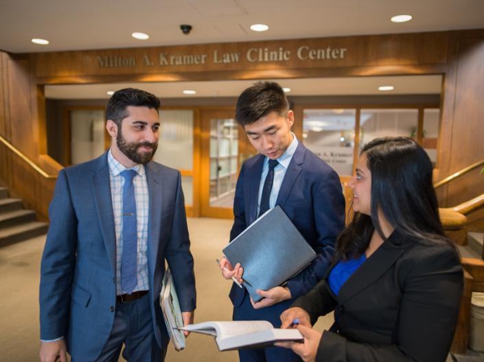 Three people in suits holding an open book and standing outside of doors that read Milton A. Kramer Law Clinic Center.