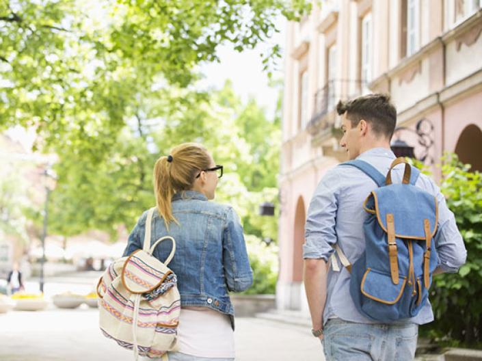 Image of two students walking away, with their backs to the camera, one wearing a jean jacket and white canvas backpack with brown and grey stripes and glasses, the other wearing a light blue shirt, jeans, and a denim fabric backpack with brown straps. They are in the sun approaching a bright green tree on their left and a beige building on their right. They are on a white path.