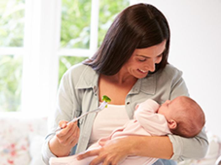 Image of mother holding baby and holding a fork with a piece of brocolli on it and smiling down at the baby, with a large window and couch with white and pink patterned cushions behind her