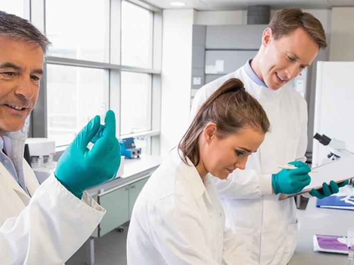 Image of three lab professionals in white lab coats and green latex gloves, the first holding up a test tube and looking at it, the second looking down at something outside the frame in front of her, the third holding a clipboard looking over the shoulder of the second and writing on the clipboard.  they are in a lab environment with bright widows to the left and 3 medium sized pieces of equipment, one a microscope, with white shelves and miscellaneous items on the right.