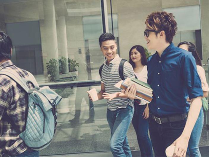 Image of five students walking in the sun in front of a large glass window.  they are wearing jeans.  two are wearing backpacks, one is holding a cup, and one, wearing sunglasses, is carrying books in one hand and a cup in the other.  The two other students are walking in the background, obscured by the three in the foreground