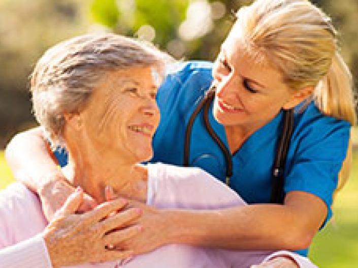 Image of health professional with blue shirt and black stethescope, with blond hair in a pigtail, with her arms around a senior in a pink blouse, with a bright sunny green field ending in a wooded area behind them