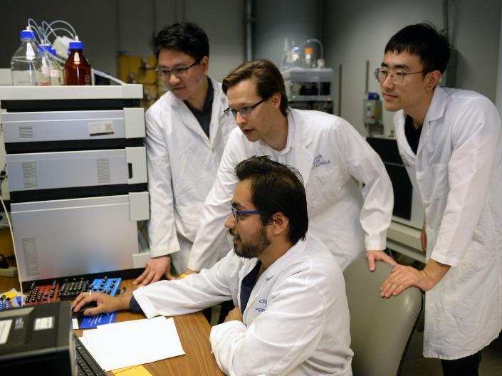 Four men in white lab coats gather around a computer.  One is sitting and the other three are standing.