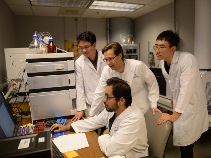 A teacher and three students wearing white lab coats gather around a computer monitor to analyze data.