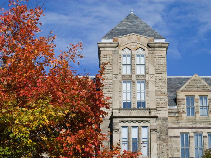 A tan brick building stands against a blue sky with a tree with red leaves in the foreground.