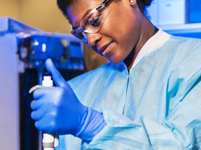 A woman wearing gloves, a medical gown, and goggles puts a sample in a test tube.