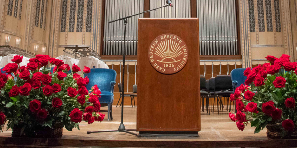 From of stage with podium center, roses on either side, and CWRU School of Medicine white coats and chairs in background.