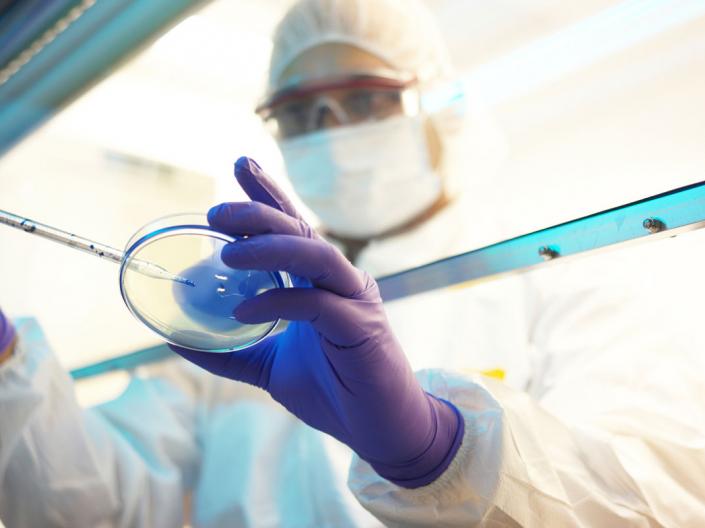 Person wearing a lab coat and face mask working in a lab with a petri dish