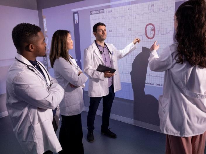 Four people in lab coats review a heartbeat graphic projected on the wall