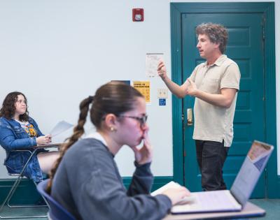 A classroom setting with a teacher standing and speaking. Two students, one focused on papers and the other on a laptop, listen attentively.
